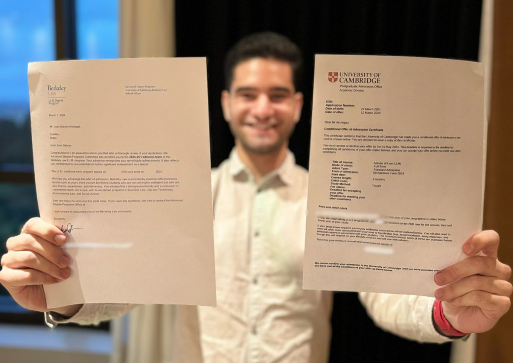 Homem segurando dois documentos no tamanho A4 perto da câmera, com o rosto ficando entre as duas folhas, que são as cartas de aprovação dos programas de Mestrado em Direito que João foi aceito.