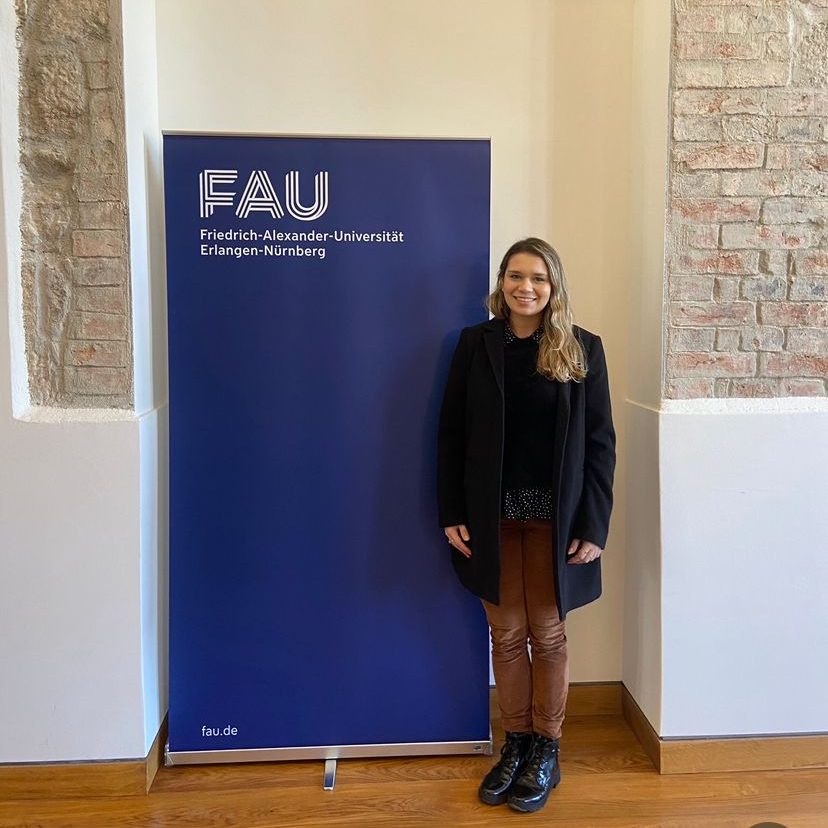 Patricia ao lado de um banner com o nome da Universidade da Alemanha FAU.