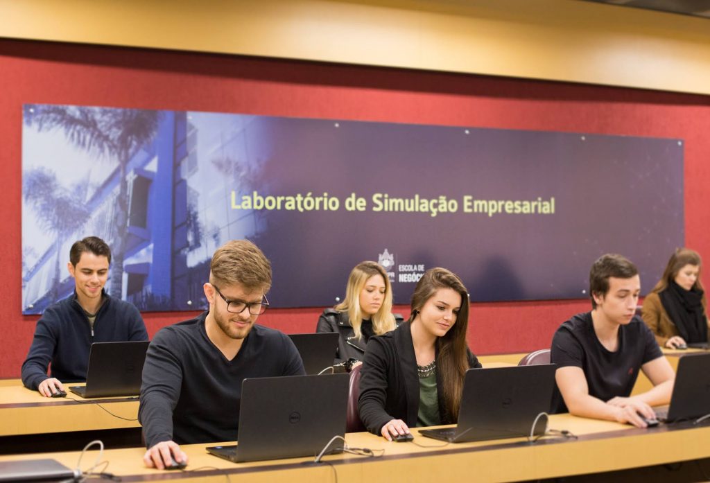 Fotografia de uma sala de aula, com três alunos aparecendo por inteiro: um homem branco, de cabelos loiros e usando óculos; uma mulher branca, de cabelos morenos longos; e uma mulher branca, de cabelos loiros longos. Os dois primeiros alunos estão na primeira fileira, olhando para notebooks. Ao fundo, a parte vermelha e amadeirada com uma placa azul na horizontal indica o local como "Laboratório de Simulação Empresarial".