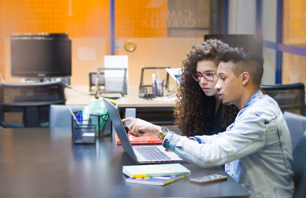 Foto caracterizando dois jovens, um rapaz e uma moça morenos, conferindo informações em um notebook em um fundo de sala de aula tecnológica.