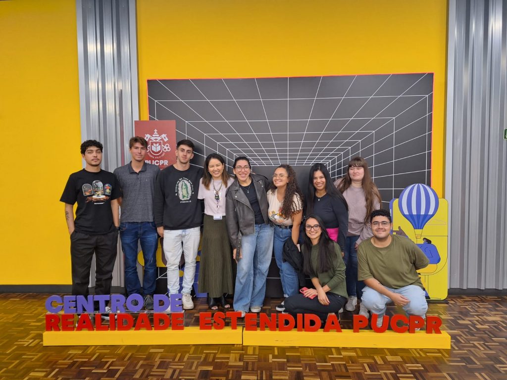 Foto da turma com uma professora ao centro e 9 alunos adultos ao redor, em frente a um banner que remete à noção de tecnologia e realidade estendida.