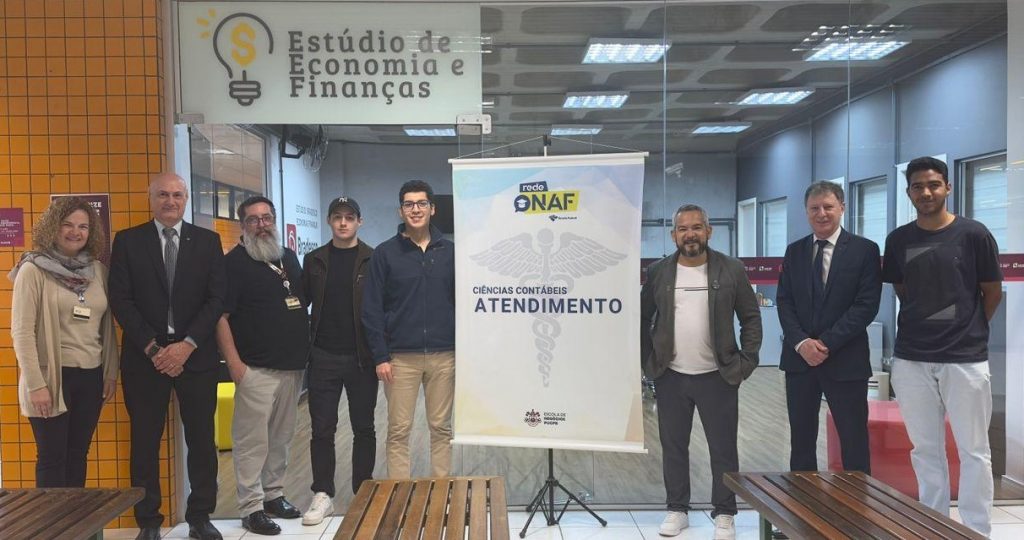 Fotografia da equipe participante do evento "Regularização do CPF", ao lado de um banner com o símbolo de Ciências Contábeis, e em frente ao Estúdio de Economia e Finanças, da Escola de Negócios da PUCPR