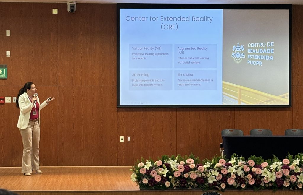 Fotografia da professora doutora Miriam Stolses Mazo, em um palco com flores na frente, apresentando um slide. O slide possui textos do lado esquerdo e uma foto com a logo do Centro de Realidade Estendida do lado direito.