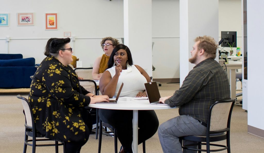 Fotografia de 3 pessoas em primeiro plano, conversando em torno de uma mesa redonda, em um ambiente claro de coworking. As pessoas são uma mulher negra, uma pessoa não-binária branca e um homem branco. Todos os três integrantes da foto são pessoas gordas. Há dois notebooks na mesa e um caderno de anotações.