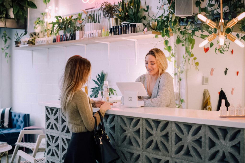 Fotografia de duas mulheres realizando uma compra. Ambas loiras e brancas, uma atrás de um balcão e com um tablete em um apoio branco, e a outra na frente, olhando para o celular. Ambiente com várias plantas, paredes de tijolos brancos e iluminação focada.