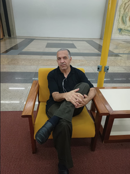 Foto de Josias, que está sentando em uma poltrona amarela com as pernas cruzadas e segurando uma das pernas.